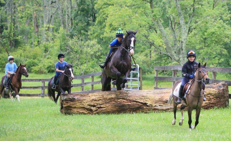 group of children on horses jumping log in field at pony camp