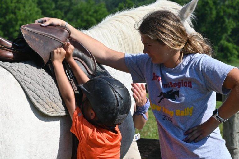 child tacking up horse at pony camp with an adult supervising