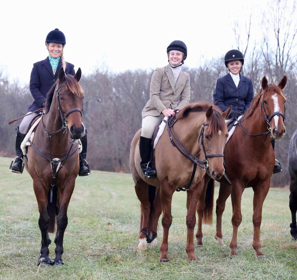 3 smiling juniors in foxhunting attire on horses in field